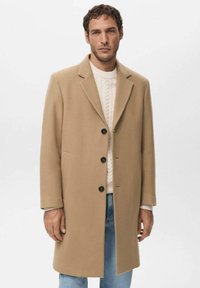 Abrigo de lana beige con cuello estructurado, tres botones negros, bolsillos laterales y superficie texturizada, llevado sobre un suéter blanco de punto.