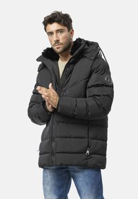 Schwarze Steppjacke mit pelzgefütterter Kapuze, Reißverschluss und gesteppter Optik; getragen über einem leichten Pullover und Jeans.