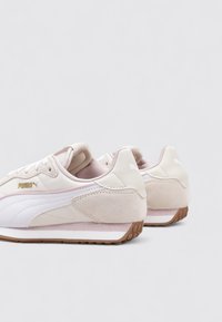 Un par de zapatillas Puma beige y blancas con suelas de goma, vistas desde la parte trasera y ligeramente inclinadas hacia un lado sobre un fondo blanco.