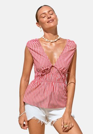 Vrouw draagt een rood-wit verticaal gestreept mouwloos topje met diepe V-hals en witte denim korte broek met rafels, sluit haar ogen en lacht.