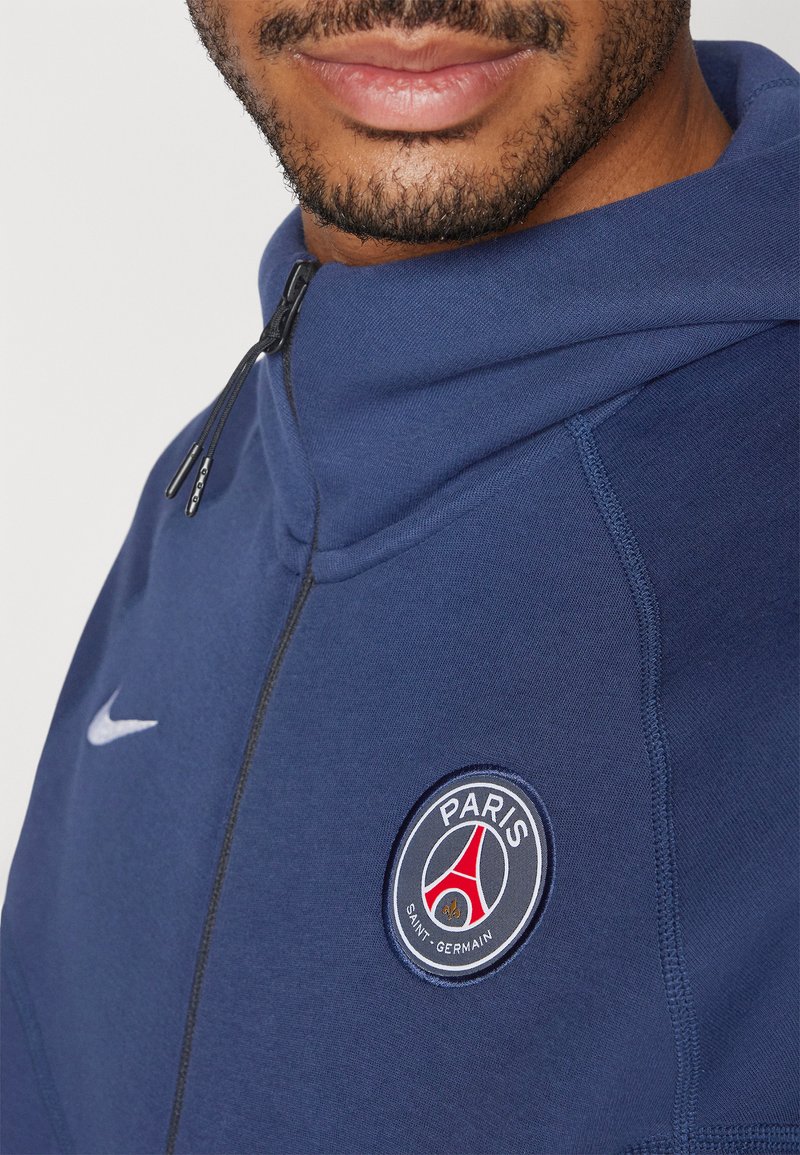 Felpa con zip di colore blu navy, con una superficie liscia, dotata di logo Nike e di un distintivo ricamato del Paris Saint-Germain sul petto.