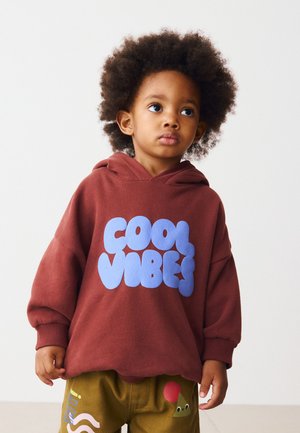Ungt barn med afro-frisure iført en mørkerød hættetrøje med teksten "COOL VIBES" og grønne bukser med farverige designs, stående mod hvid baggrund.