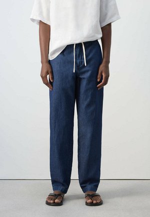 Persona que lleva pantalones vaqueros sueltos de color azul oscuro con cintura de cordón, camiseta blanca de manga corta y chanclas negras, de pie sobre un suelo gris.