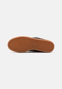 Black sneaker with a textured gum rubber sole featuring a patterned grip. Notable logo detail is embossed on the midsole.