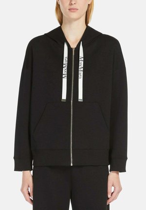 Donna che indossa una felpa nera con zip e coulisse bianche con etichetta "Max Mara" e pantaloni neri abbinati, in piedi su uno sfondo bianco.