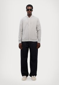 Grå hettejakke med glidelås og snorer, kombinert med mørkeblå vide jeans og beige sneakers. Modell med solbriller som står foran en ensfarget bakgrunn.
