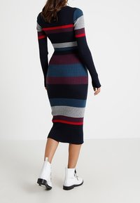 Hedvábná pletená šaty s dlouhými rukávy se horizontálními pruhy v námořnické modři, šedé, červené a tyrkysové barvě. Nosené s bílými kotníkovými botami.