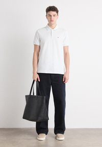 Hvid kortærmet polo skjorte med et lille rødt logo, parret med mørkeblå brede bukser og beige sneakers, mens der holdes en sort tote taske.