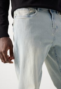 Light blue denim jeans with a faded finish, five-pocket design, and subtle texture. A hand is positioned near the waistband.