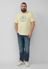 Geel T-shirt met korte mouwen en zwart ronde grafische tekst, gecombineerd met blauwe jeans en witte sneakers. Casual pasvorm, gemaakt van katoen.