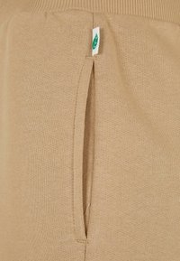 Beige strikkegenser med en sidelomme og et grønt bladmærke. Stoffet har en jevn tekstur og ribbestrikket halslinning.