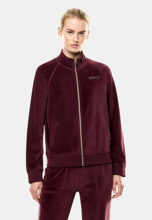 Donna che indossa una giacca di velluto color borgogna con cerniera e pantaloni abbinati, in piedi davanti a uno sfondo chiaro e uniforme.