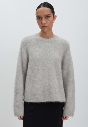 Femme aux cheveux bruns tirés en arrière, portant un pull en laine épaisse gris clair et une jupe noire, se tenant contre un fond gris clair uni.