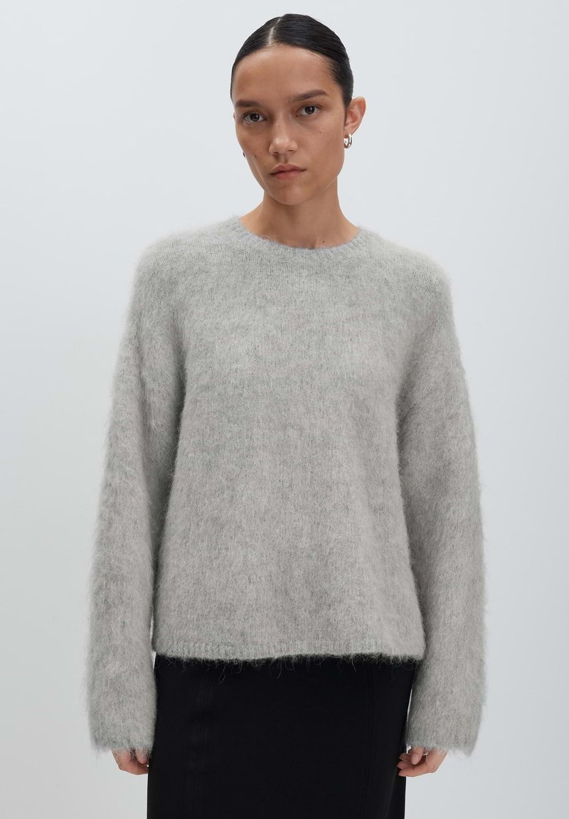 Vrouw met donker haar dat naar achteren is vastgebonden, draagt een lichtgrijze fuzzy trui en een zwarte rok, terwijl ze tegen een effen lichtgrijze achtergrond staat.