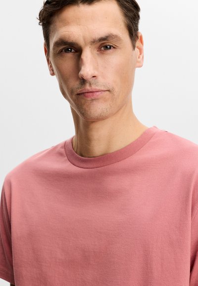 Jeune homme aux cheveux courts et foncés portant un t-shirt rose uni à col rond, regardant droit devant avec une expression neutre.