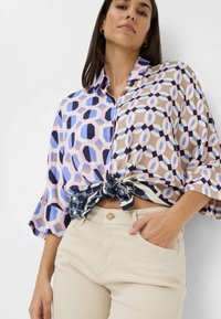 Patroon blouse in pastelkleuren, met geometrische vormen, lange mouwen en een geknoopt voorpand. Gecombineerd met lichtbeige broek.