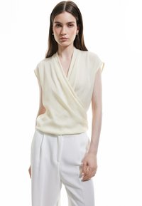 Lyscremet bluse med indpakningsdesign, korte ærmer og blød drapering. Parret med hvide, højtaljede bukser, der fremhæver et rent look.