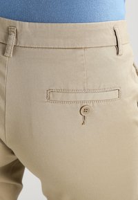 Närbild av beige byxor med en bakficka med knappstängning och en del av en blå skjorta instoppad.