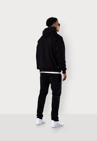 Sweatshirt à capuche noire avec une coupe ample, associé à un jean noir et des baskets blanches. Le design présente une étiquette sur l'ourlet.