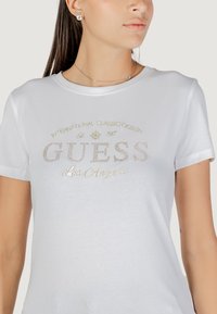 T-shirt bianca in cotone con maniche corte, scollo rotondo e dettagli in strass dorati, che riportano la scritta "GUESS Los Angeles" sopra elementi decorativi.