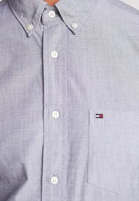 Vaaleanvioletti napillinen paita, jossa on teksturoitu kangas, kaulus ja tasku, jossa pienenpieni kirjailtu logo punaisena, valkoisena ja sinisenä.