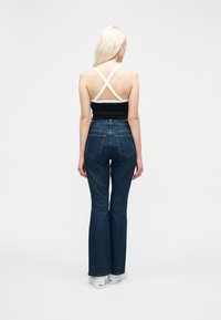 Γυναίκα που στέκεται πλάτη, φορώντας σκούρα μπλε τζιν, μαύρο τοπ με άσπρα λουριά σε σχήμα σταυρού και άσπρα παπούτσια με πλατφόρμα σε λευκό φόντο.