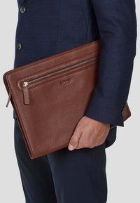 Brun lær dokumentsak med en teksturert finish, med glidelås på toppen og en utvendig lomme, holdt av en hånd i et marineblått dress.