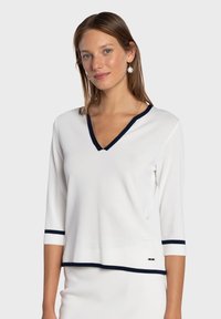 Pull-over blanc avec col en V et manches trois-quarts. Présente un bord bleu marine au niveau de l'encolure et des poignets. Confectionné dans un matériau doux.