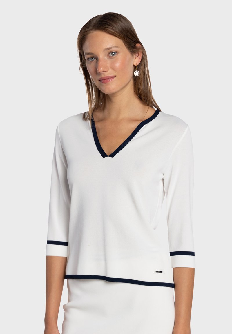 Pull-over blanc avec col en V et manches trois-quarts. Présente un bord bleu marine au niveau de l'encolure et des poignets. Confectionné dans un matériau doux.