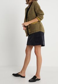 Nainen, joka on pukeutunut vihreään neulepaitaan, mustaan toppiin, tummaan samettihameeseen ja mustiin slip-on loafer-kenkiin, seisoo vaaleaa valkoista taustaa vasten.