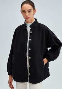 Zwarte denimjack met een knoopsluiting aan de voorkant, hoge kraag en een loose fit. Heeft twee zijzakken en zilveren knopen. Lichtjes gestructureerde stof.