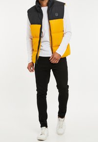 Veste matelassée à blocs de couleur avec une partie supérieure noire et une partie inférieure jaune vif. Conception légère avec fermeture éclair, col montant et écusson logo.