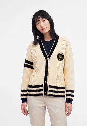 Femme portant un cardigan crème avec des rayures noires, des boutons dorés, un écusson sur la poitrine et un pantalon beige sur un fond blanc uni.