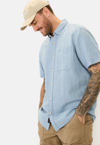Lichtblauw korte mouwen button-up shirt met een borstzak, gemaakt van textuurstof. Gecombineerd met beige shorts en een tan pet.