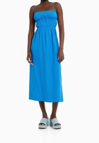 Žena v jasně modrých midi šatech s tenkými ramínky a nařaseným živůtkem, doplněná světle modrými sandály na platformě s otevřenou špičkou.