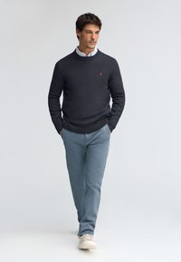 Dunkelgrauer Strickpullover mit geripptem Ausschnitt, kombiniert mit hellblauen Hosen und beigen Sneakers, getragen von einem Mann vor neutralem Hintergrund.