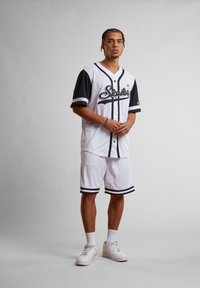 Maillot de baseball blanc avec des accents noirs et le logo "Starter", associé à un short blanc avec des rayures noires, porté avec des baskets blanches.