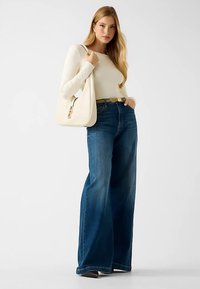 Jean en denim bleu à jambes larges avec un haut à manches longues crème clair. Le modèle tient un sac à main blanc. Texture lisse, design décontracté.