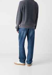 Mann in einem grauen Strickpullover, blauen Jeans und weißen Sneakers, der vor einem einfarbigen weißen Hintergrund steht und in die entgegengesetzte Richtung schaut.