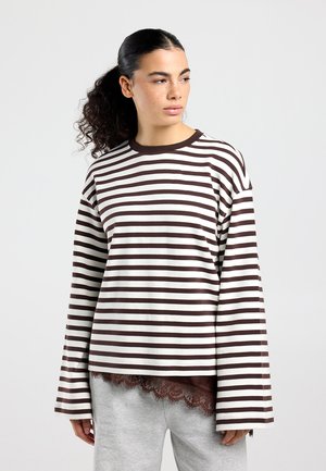 Giovane donna con capelli ricci raccolti in una coda alta, che indossa una maglia a maniche lunghe a righe marroni e bianche e pantaloni grigio chiaro, che guarda verso sinistra.