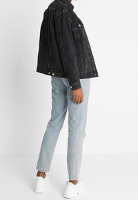 Person klädd i en svart denimjacka, ljusblå jeans och vita chunky sneakers som står mot en vit bakgrund.