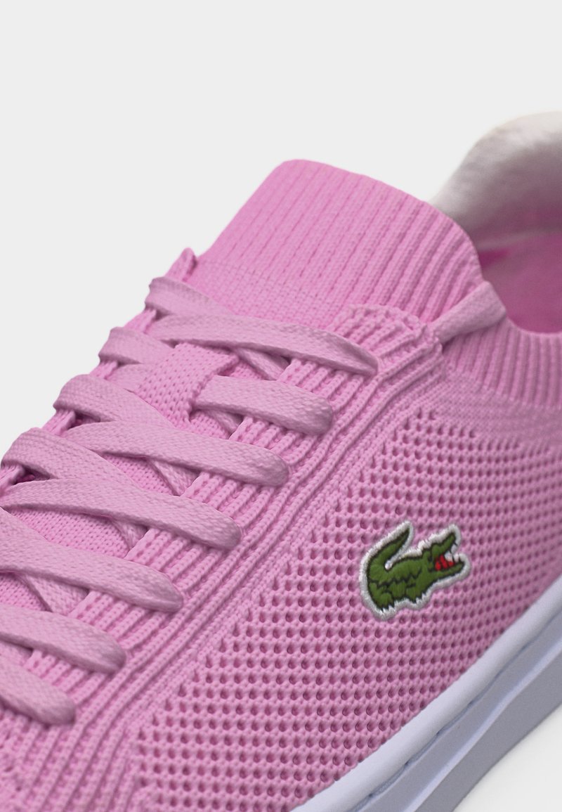 Rosa gestrickter Sneaker mit Schnürsenkeln, elastischem Knöchelkragen und grünem Krokodil-Logo an der Seite, vor weißem Hintergrund.