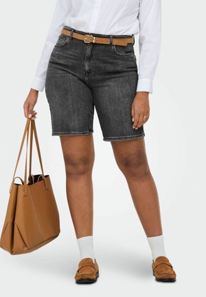 Kvinne iført mørke denimshorts, hvit skjorte, brunt belte, hvite sokker, brune loafers, og holder en stor brun handlenett ved siden av seg.