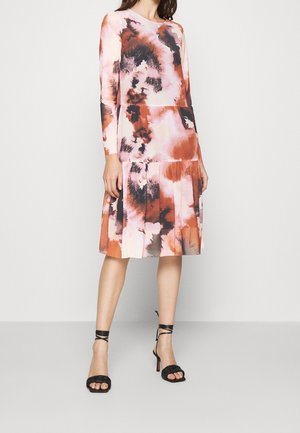 Femme portant une robe à manches longues, longueur genoux, avec un motif abstrait rose, orange et noir, associée à des sandales noires à talons hauts et lanières.