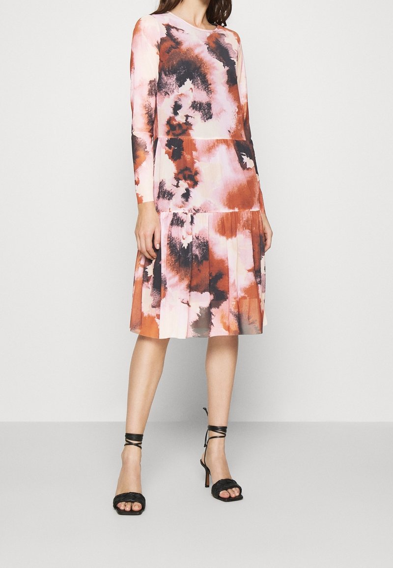 Femme portant une robe à manches longues, longueur genoux, avec un motif abstrait rose, orange et noir, associée à des sandales noires à talons hauts et lanières.