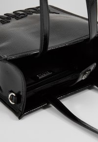 Czarna torebka z lakierowanej skóry z podwójnymi uchwytami, otwarta klapa ukazująca wnętrze z kieszenią na zamek i wytłoczonym logo marki na górze.