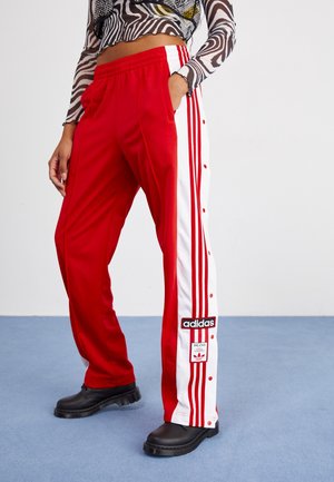 Osoba nosząca czerwone dresowe spodnie Adidas z białymi panelami bocznymi i czerwonymi paskami, w połączeniu z czarnymi butami i wzorzystą, długorękawową bluzką.