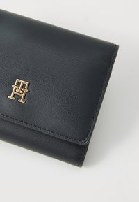 Portofel din piele neagră cu suprafață texturată, logo din metal auriu pe față și închidere prin pliere. Detaliu de cusături vizibile.