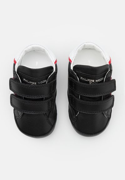 Sapatos de bebé em couro preto com detalhes brancos e vermelhos, apresentando duas correias de Velcro e um design de ponta arredondada. Textura suave.