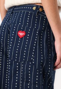 Laivastonsiniset housut, joissa valkoiset raidat ja pienet sydänkuviot, takataskussa punaista sydämen muotoista kangasmerkkiä, jossa lukee "Carhartt WIP".